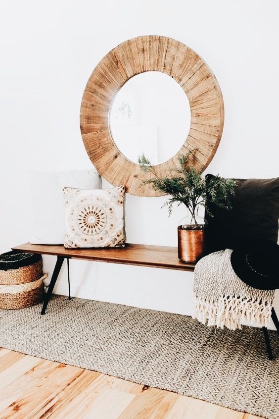 a simple boho entryway with a wooden bench, a woven rug, a basket, a wood clad mirror and pillows