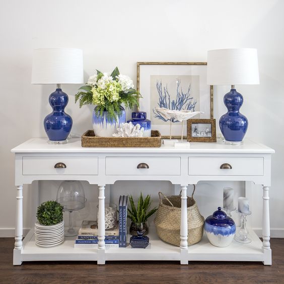 a white vintage console with bold blue lamps and vases, plants in pots, baskets and corals