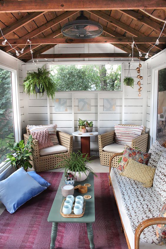 a boho screened porch with lights, hanging potted greenery, rattan and wooden furniture, a colorful rug