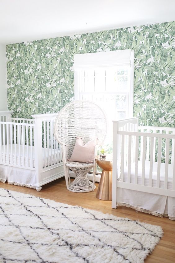 a light-filled shared nursery with a palm leaf print wall, white cribs and a tropical-inspired chair
