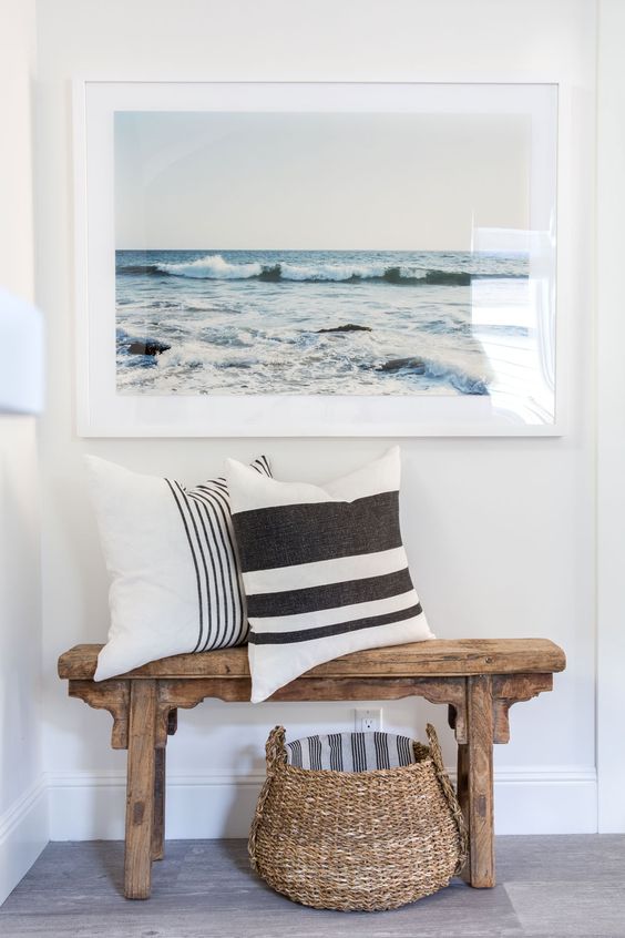 a simple beachy entryway with a wooden bench, a basket, a sea artwork and striped pillows