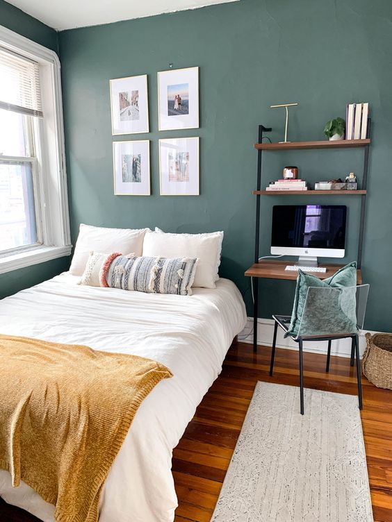 a dark green guest bedroom with a bed and neutral bedding, open shelves with a desk, a small chair and a gallery wall