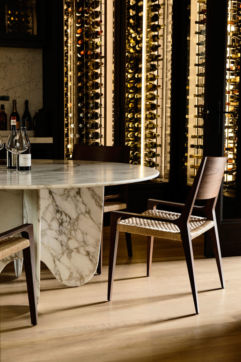 A small dining space features a marble table, woven chairs and a large lit up glass wine cooler