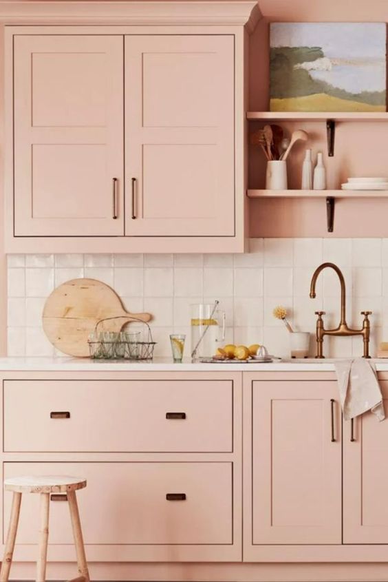 a blush kitchen with a white square tile backsplasj and white countertops, black handles and a brass faucet