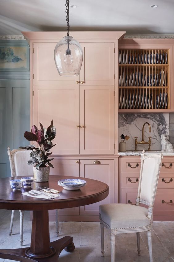a chic pink kitchen with shaker style cabinetry, a stained round table, white chairs, a pendant lamp and a white stone backsplash