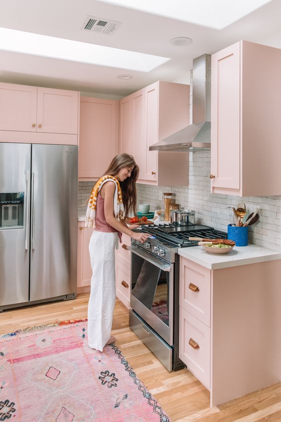 a light pink kitchen with shaker cabinets, white stone countertops, a white brick backsplash and brass handles