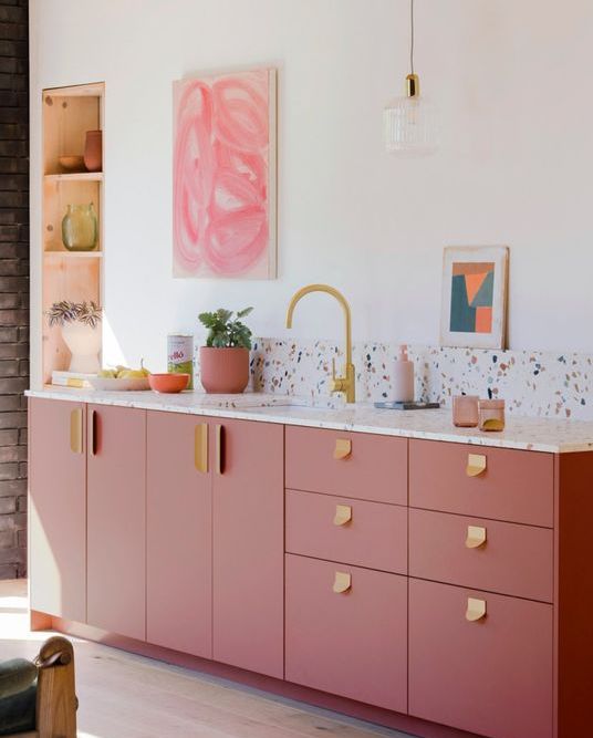 a modern and stylish pink kitchen with a terrazzo backsplash and countertops, gold hardware and a faucet, a niche for storage