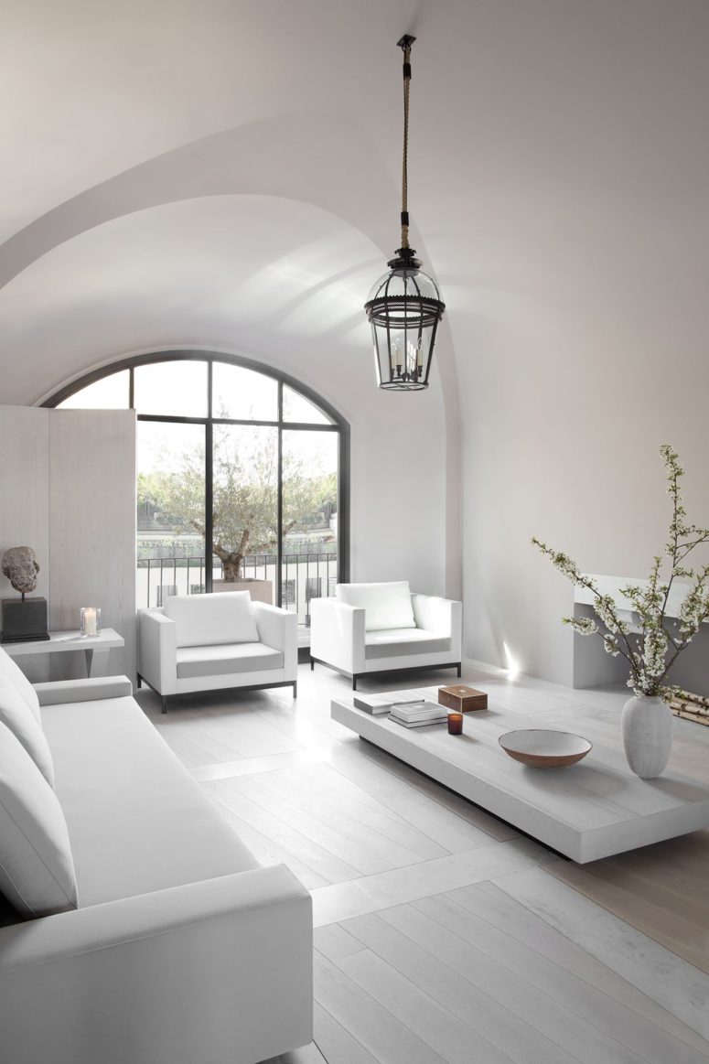 The living room is a light filled space with white furniture and much light coming through an arched window The living room is a light-filled space with white furniture and much light coming through an arched window