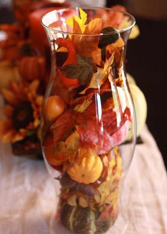 a simple and cute display of centerpiecce of a sheer vase filled with fake fall leaves and pumpkins