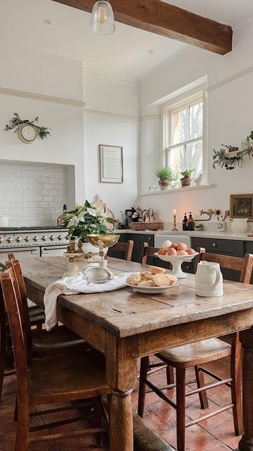 a beautiful farmhouse kitchen with navy lower cabinets, a cooker and a built-in hood, a stained table and chairs and some greenery