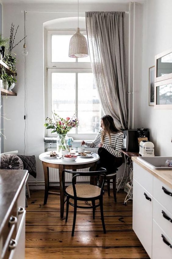 a little Scandinavian kitchen with white cabinets and a small eating zone at the window, with a round table and cane chairs
