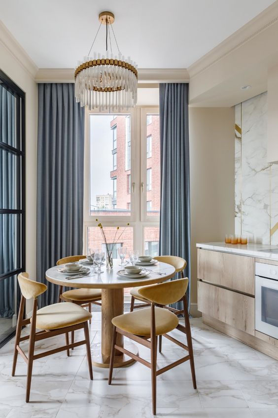 a small and pretty eat-in kitchen with stained lower cabinets and a marble backsplash, a light-stained dining set and a chic crystal chandelier