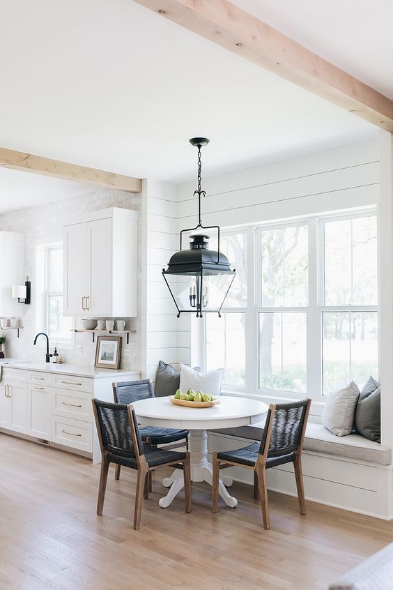 a stylish farmhouse kitchen with planked walls and shaker cabinets, a windowsill bench and a round table plus black chairs