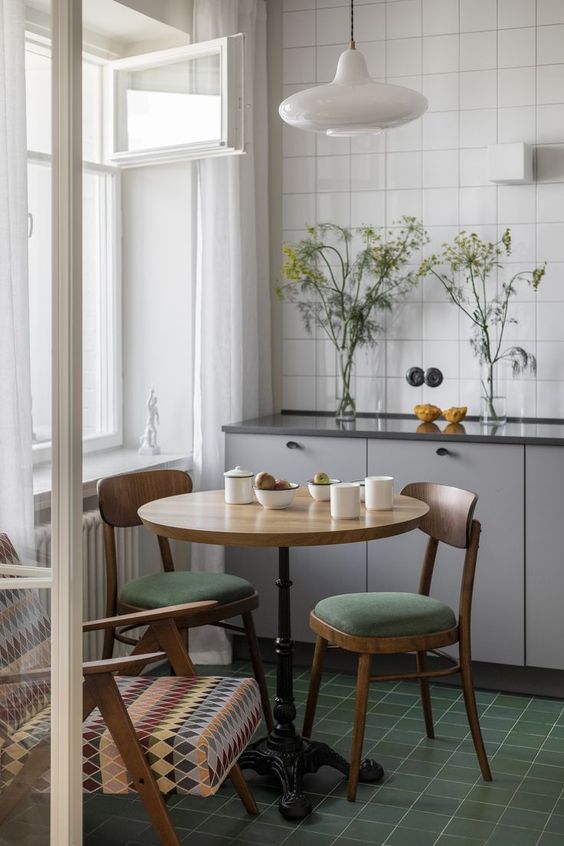 a tiny Scandi eat-in kitchen with lower grey cabinets, a white square tile backsplash, a vintage table and matching chairs plus a green tile floor