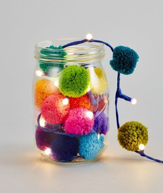 a jar with colorful pompoms and lights is a lovely decoration and table lamp for Christmas that can be DIYed