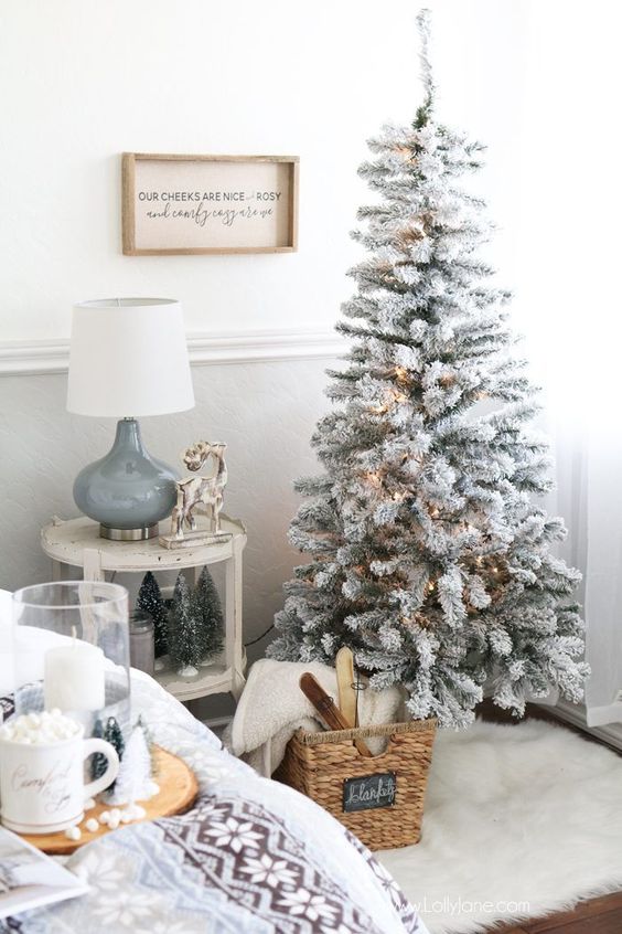 a cozy neutral space with a flocked Christmas tree with lights, a basket with fur and skis and tinsel trees
