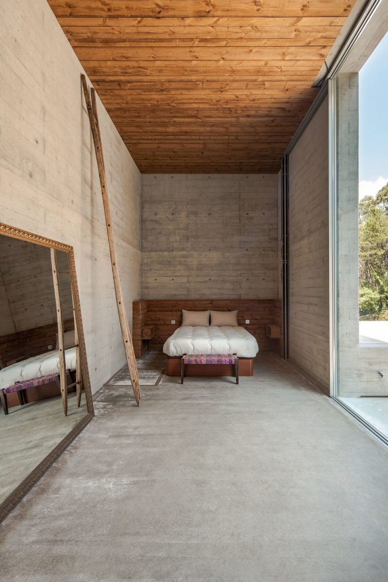 Wood panels and furniture add coziness to this industrial bedroom done with concrete, there's a large mirror and a colorful footrest