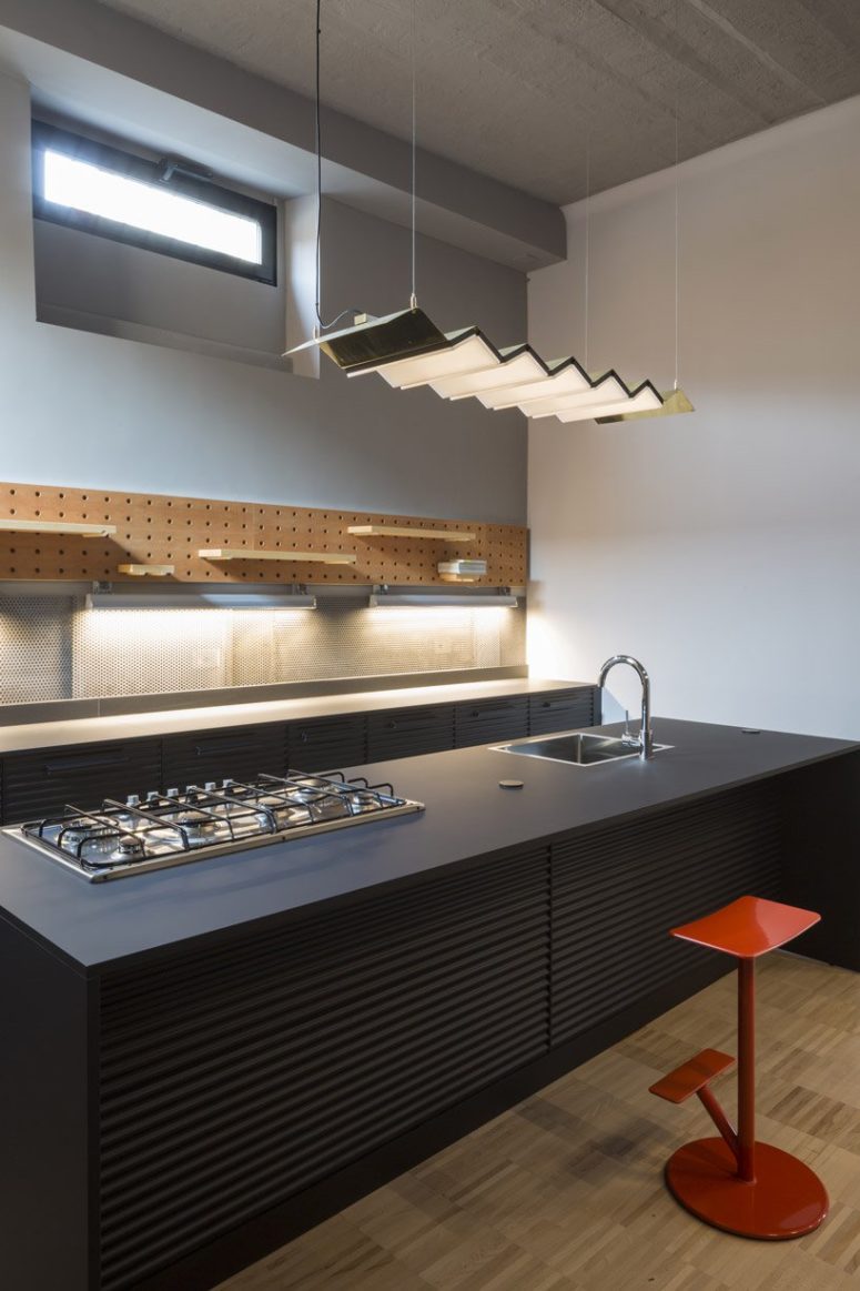 The kitchen is done with black metal cabinets, a pegboard with various shelves attached and a pendant lamp