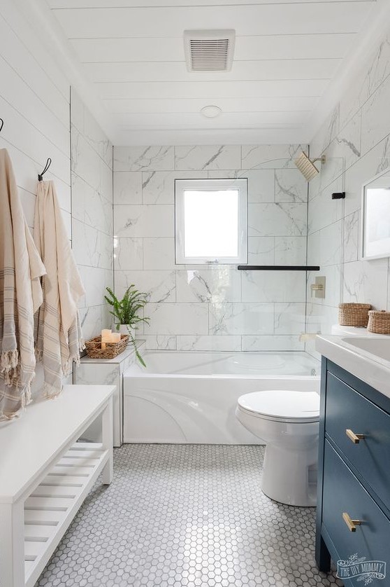 a beach bathroom with white marble large format and hex tiles, a navy vanity, a white bench and some baskets and towels