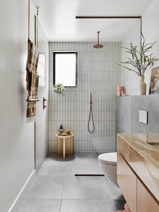 a beautiful grey bathroom with skinny and large scale tiles, a floating vanity, a shower space, a wooden stool and copper fixtures