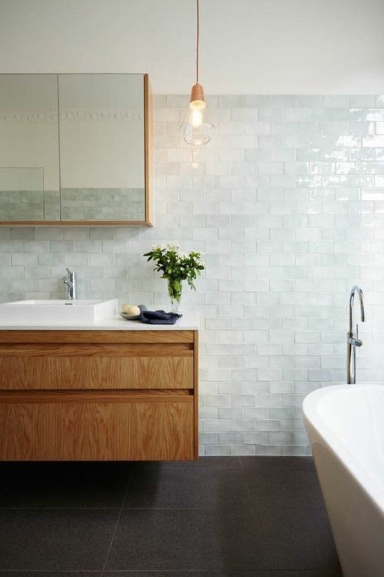 a charming coastal bathroom clad with very pale green glazed tiles, a tub, a wooden vanity and a pendant lamp