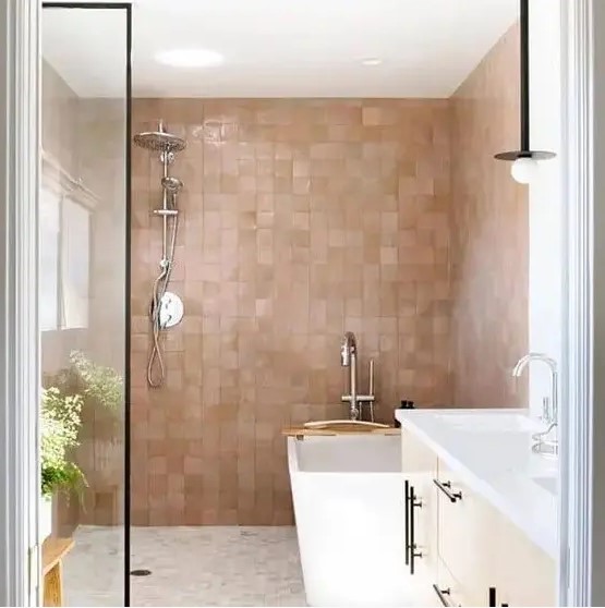 a chic bathroom with white furniture and appliances and lovely pink and tan zellige tiles covering the walls in the bathing space and giving it a soft and warming glow