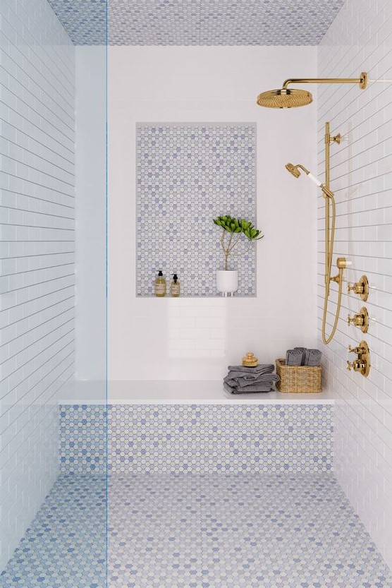 a coastal shower space done with white and blue tiles, a niche, a bench, some towels and gold fixtures