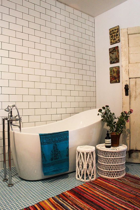 a cozy small bathroom clad with small scale blue and white subway tiles, with a tub, a gallery wall, side tables and colorful textiles