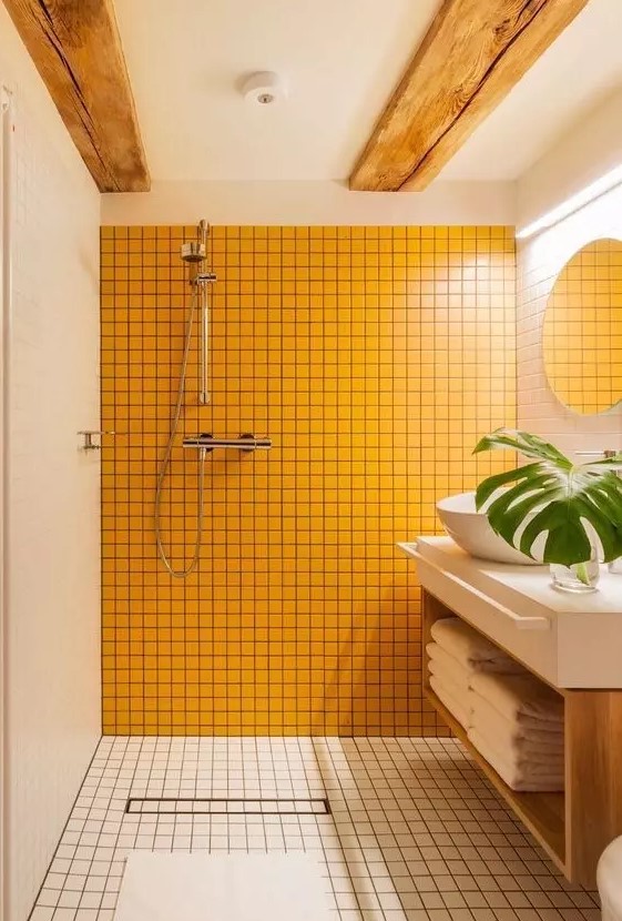 a laconic modern bathroom clad with yellow and neutral square tiles, a floating vanity with a bowl sink, wooden beams and a round mirror
