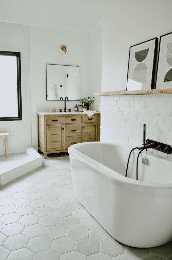 a modern eclectic bathroom with a hex tile floor, an oval tub, a wooden vanity, black fixtures and a ledge with artworks