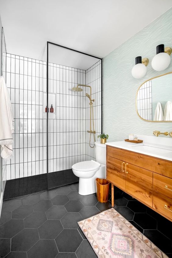 a modern farmhouse bathroom with black hex and white skinny tiles, a shower space, a timber vanity, a round mirror, gold fixtures