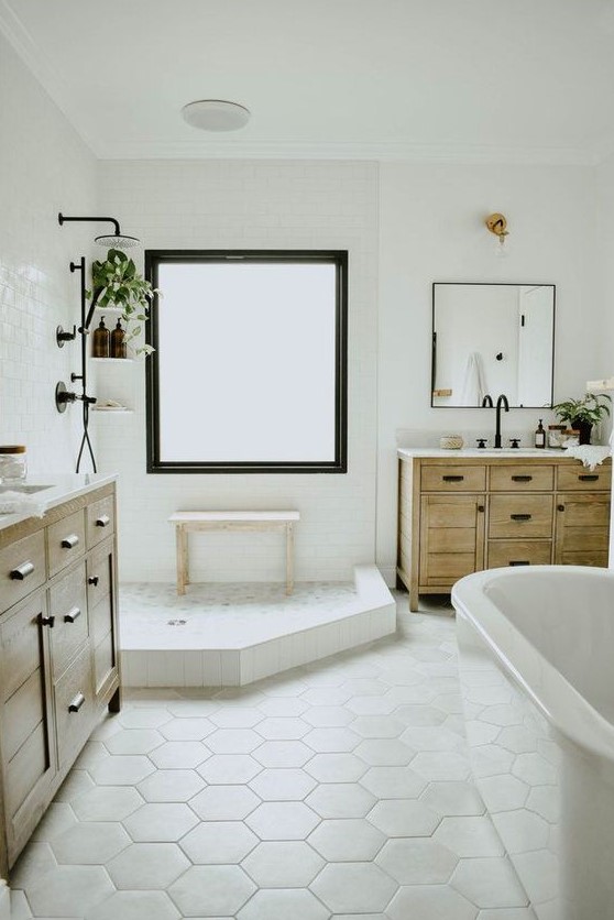 a neutral farmhouse bathroom with a two wooden vanities, an oval tub, a shower space with a large window with frosted glass