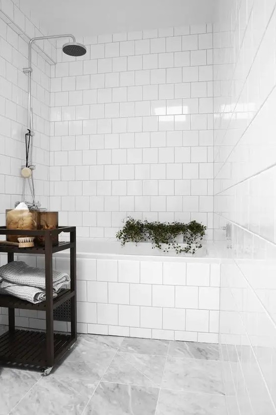 a simple white bathroom with white square tiles and a grey marble tile floor, a black shelving unit, some greenery and decor