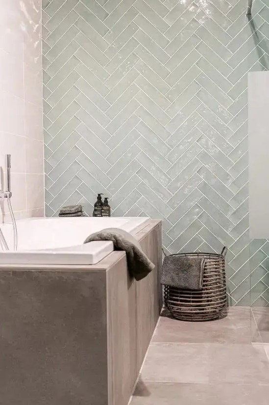 a unique contemporary bathroom clad with taupe tiles and glossy mint chevron ones, with a basket for storage and some neutral textiles