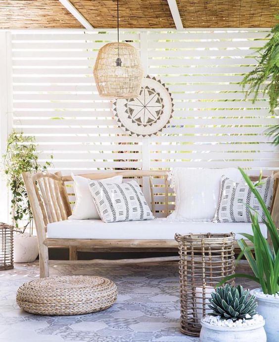 a welcoming tropical patio with a rustic wooden bench with pillows, a wicker lamp, a woven ottoman and a rattan candle holder