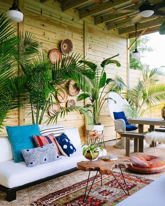 a tropical boho patio with potted plants, a white sofa, boho rugs and an ottoman, a living edge table and decorative baskets on the wall
