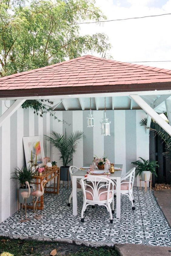 một hiên nhiệt đới với một không gian ăn uống được thực hiện với những chiếc ghế mây trắng và hồng, hồng hạc, một bàn mây và những chậu cây một hiên nhiệt đới với một không gian ăn uống được thực hiện với những chiếc ghế mây trắng và hồng, hồng hạc, một bàn mây và những chậu cây