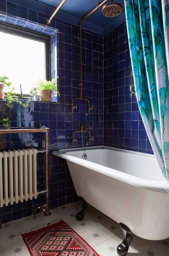 a bold bathroom with electric blue tiles, a clawfoot bathtub, a modern radiator, a bold boho rug and some potted plants