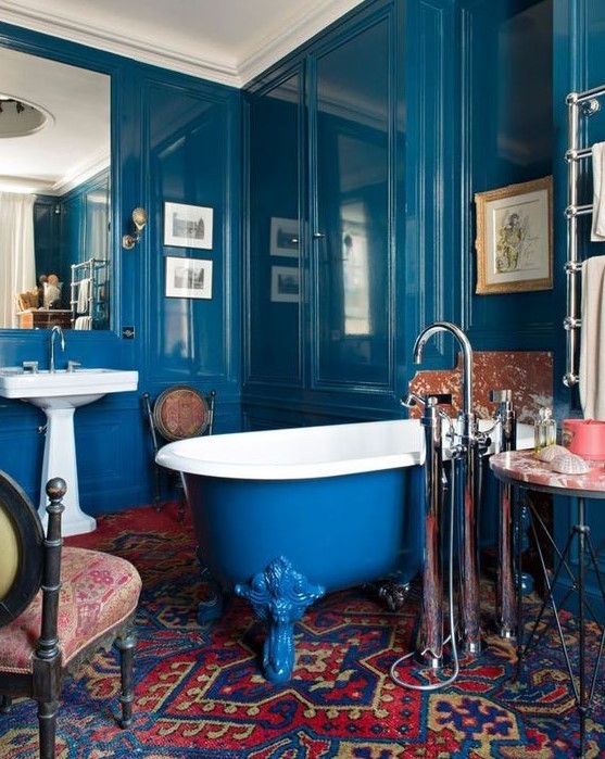 a bold bathroom with shiny paneled blue walls, a bold blue and red printed rug, a blue clawfoot bathtub and vintage furniture