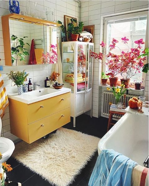 a bold eclectic bathroom with a yellow vanity, a mirror cabineta, a tub, a fluffy rug, potted blooms and greenery