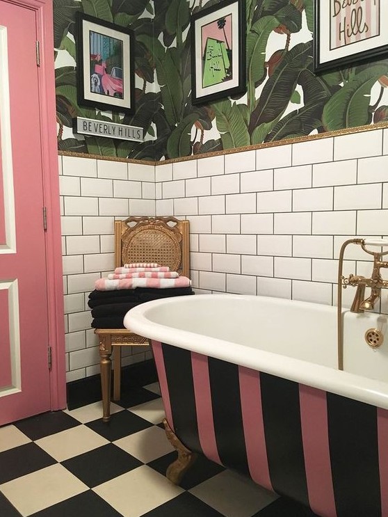 a brigth bathroom with white subway tiles, tropical leaf wallpaper, a striped bathtub, a pink door and a checked floor plus pop artworks