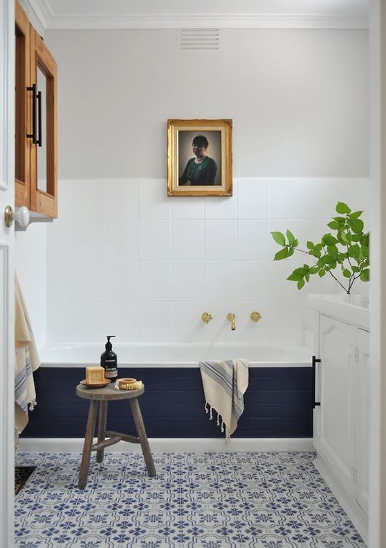 a chic eclectic bathroom with white square tiles and bold blue and white printed ones, a tub clad with navy tiles, a white cabinet and artwork