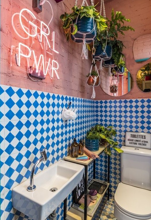 a maximalist bathroom with pink brick walls, blue and white checked tiles, a console table, potted greenery, a neon sign and mirrors