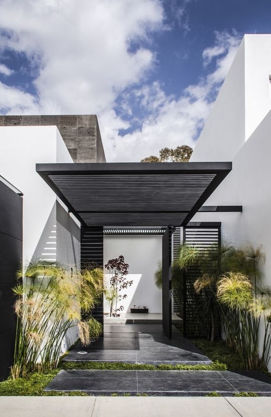 a modern front yard clad with dark tiles, with grass and tall plants and with a planked roof over the porch is very stylish and bold