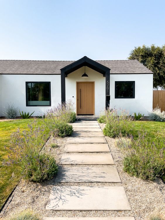 a modern to Scandinavian front yard with a stone path, some simple blooms lining it up and a green lawn is cool and chic