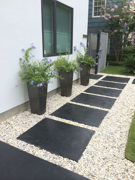 a stylish modern front yard with gravel and oversized black tiles, tall black planters with greenery and blooms