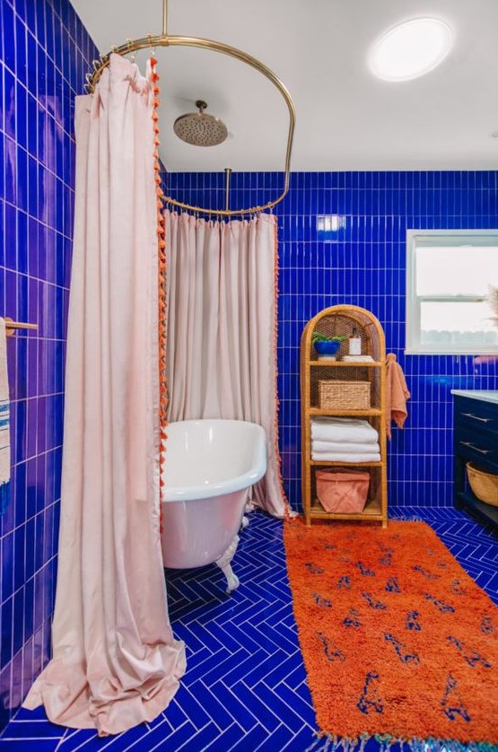 a super vibrant maximalist bathroom clad with electric blue tiles, a vintage tub with a large curtain, an orange rug, a rattan shelf and a black vanity