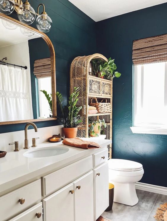 a moody eclectic bathroom with soot walls, a white vanity, a rattan shelving unit, white appliances and some potted greenery