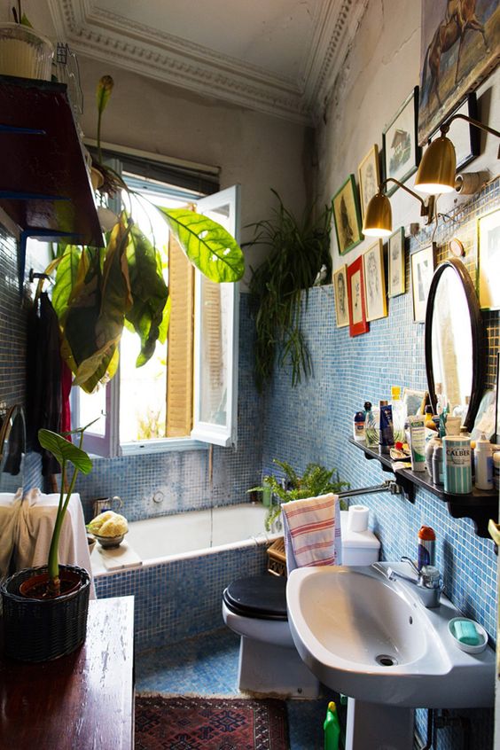 an eclectic bathroom clad with light blue tile, a tub with a window, a gallery wall, a sink and some greenery