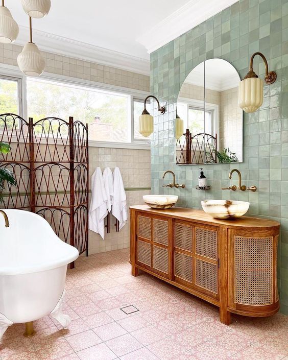 an eclectic bathroom clad with neutral and green tile, pink printed tile on the floor, a space divider, a cane vanity, a tub and some vintage lamps